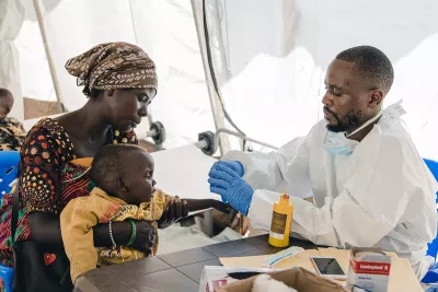 Woman holds boy as he receives treatment from health official. 