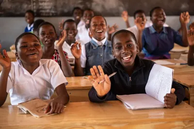 Teenage schoolgirls seated at wooden desks in a classroom, raising their hands and holding notebooks and pens.