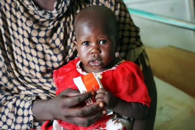 A child wearing a red dress sitting on an adult's lap is holding a RUTF sachet. 
