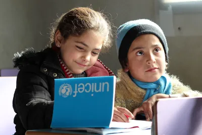 Two children sitting at a desk in a classroom, engaged in writing or drawing activities, with one of them holding a notebook with the UNICEF logo on.
