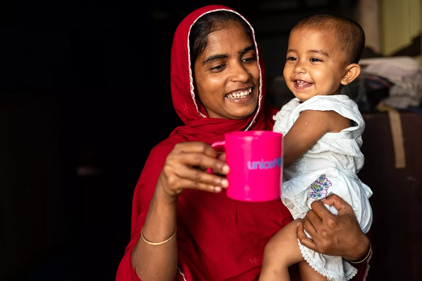 A woman smiles while holding a baby with her left arm and a mug with her right hand. 
