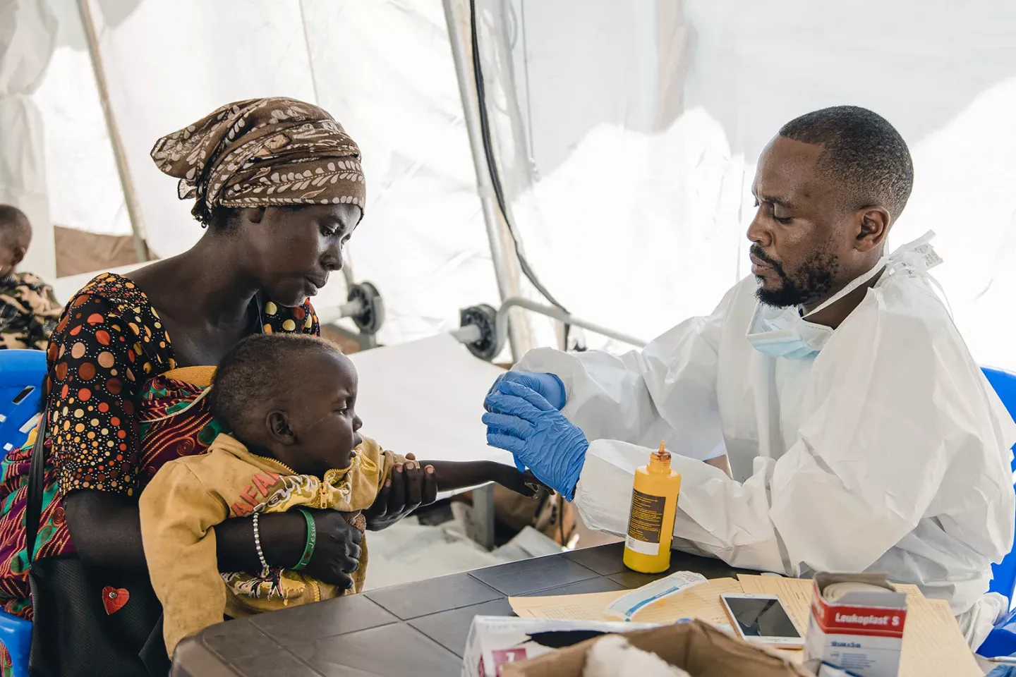 Woman holds boy as he receives treatment from health official. 