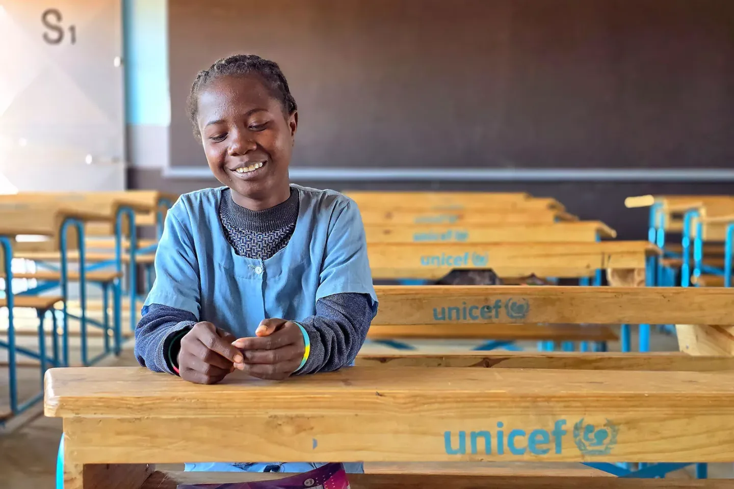 A girl smiles while sitting at a school desk in a classroom. 