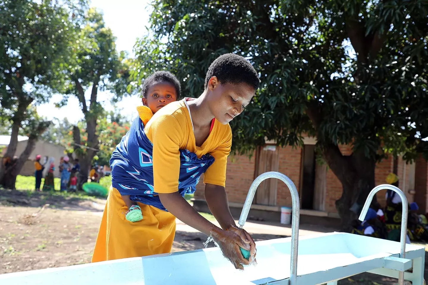 A woman washes her hands at a sink holding a baby on her back. 