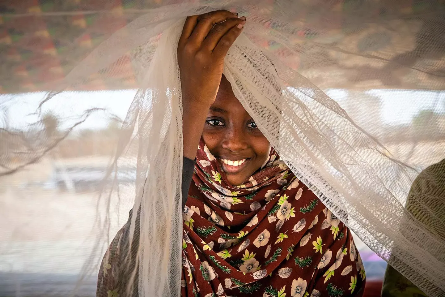 A girl smiles while lifts a mosquito net to uncover her face. 