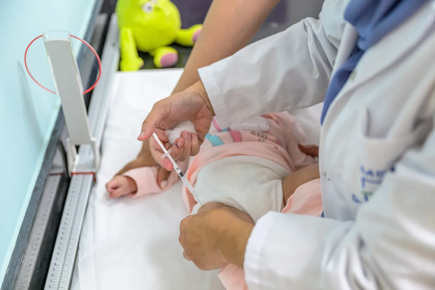 A healthcare professional in a white lab coat administers an injection into an infant&rsquo;s thigh as the baby lies on an examination table.