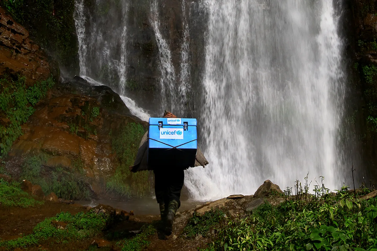 With a waterfall in the background, a porter carries UNICEF-provided vaccines on difficult terrains on the way to a vaccination campaign at the Barpak Village Development Committee (VDC) health post in Gorkha District, the epicentre of the earthquake in Nepal in 2015.