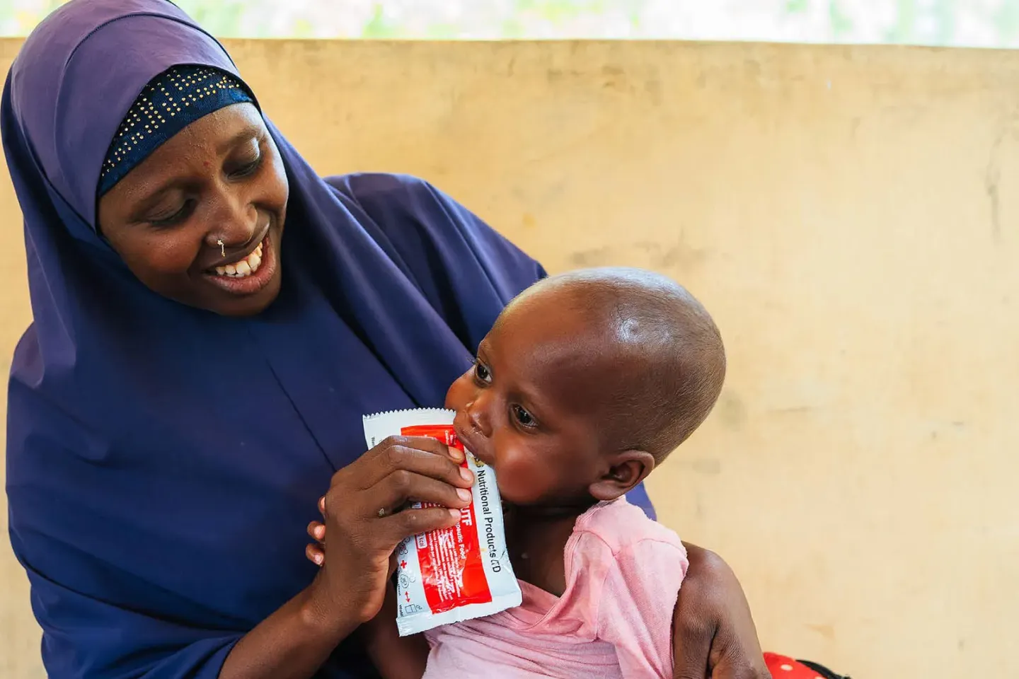 Sitting on the lap of a woman, a child eats ready-to-use therapeutic food (RUTF) from a sachet 