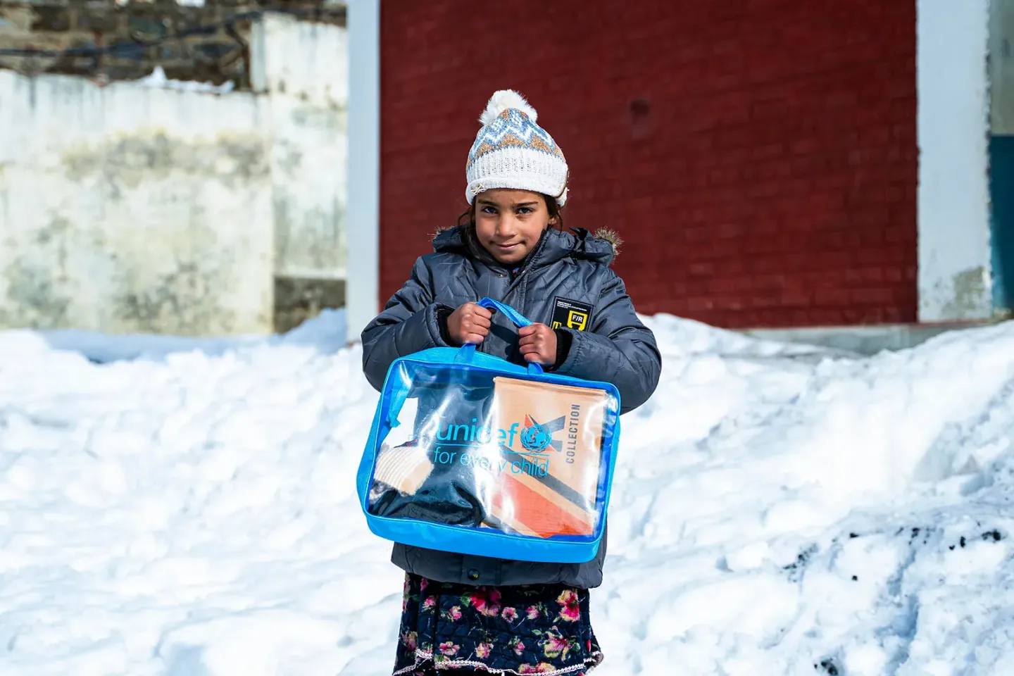 A girl standing outdoors in snow, wearing a winter coat and knit hat, holding a UNICEF-branded bag containing school supplies.