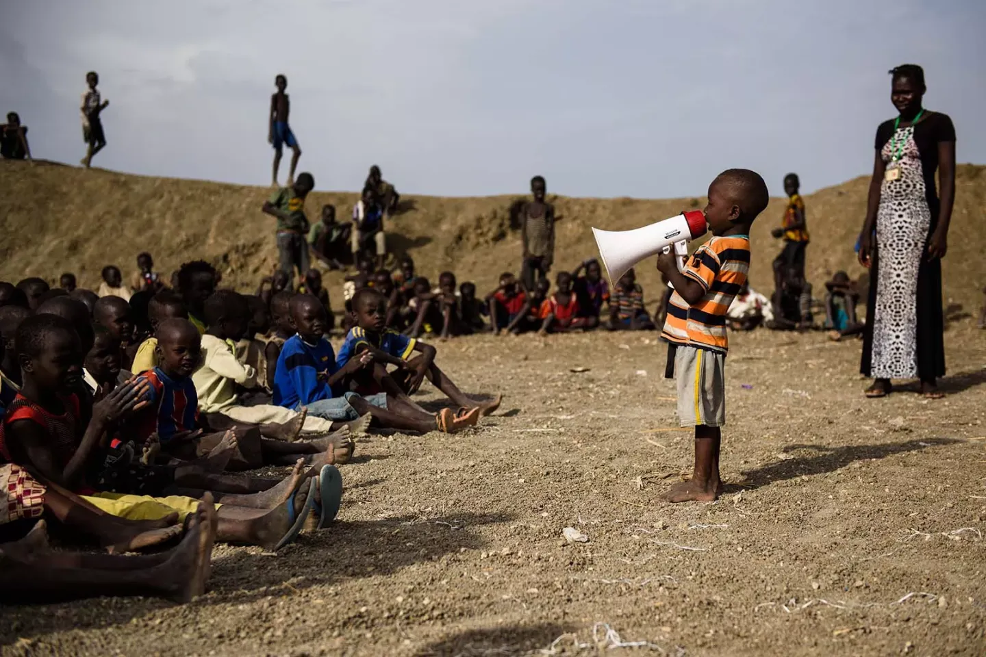 Outdoors, a boy sings into a megaphone for a group of children sitting on the ground and being watched by a woman standing beside them.