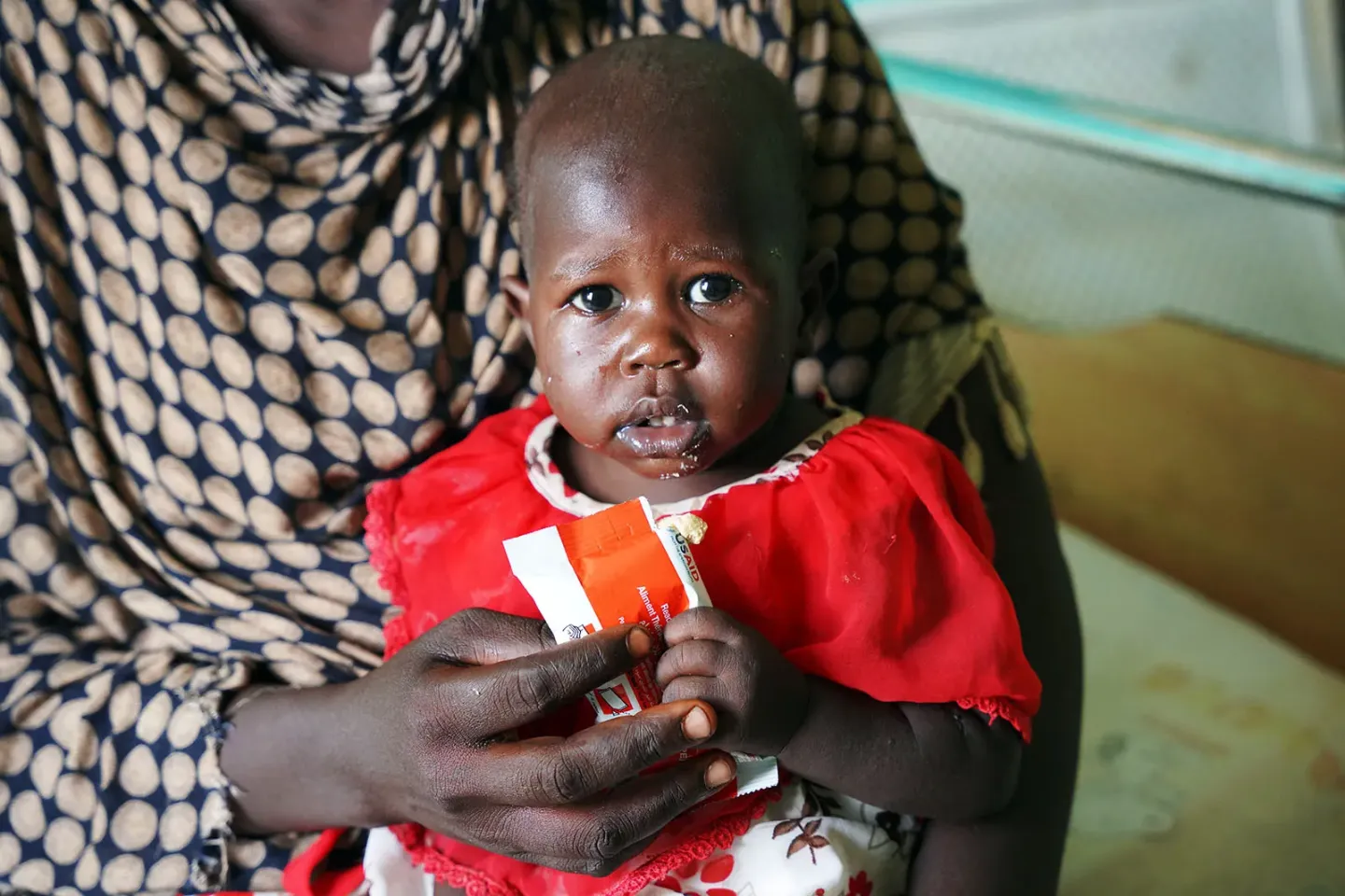 A child wearing a red dress sitting on an adult's lap is holding a RUTF sachet. 
