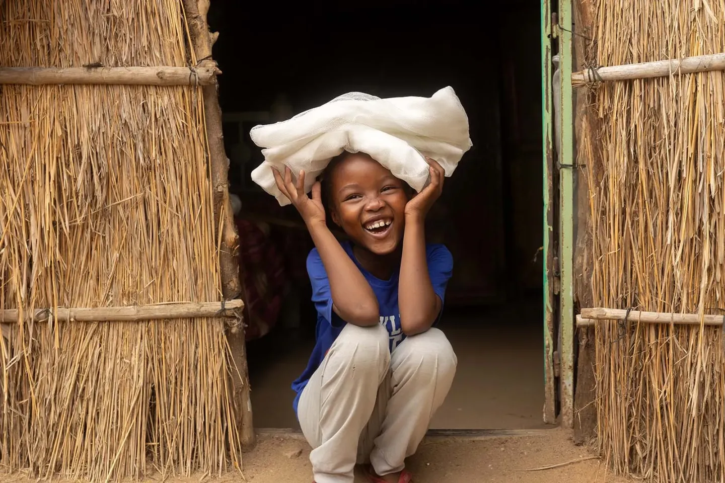 A child smiles while sitting on the ground in a doorway made of woven straw or thatch and holding a white cloth above her head. 