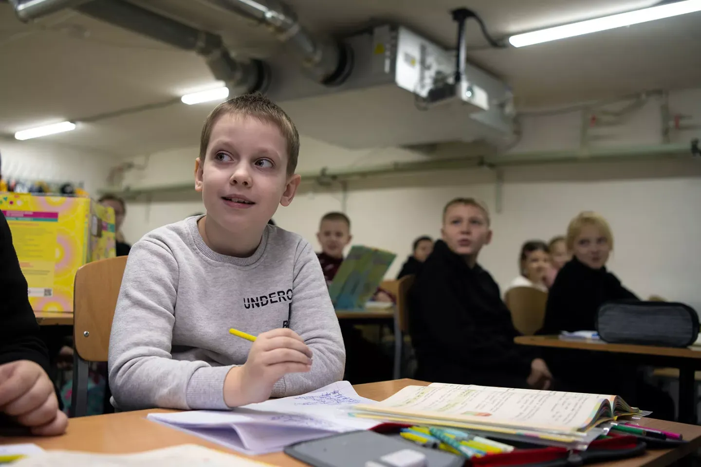A boy holding a pen sits at a desk in a classroom, where there are other children sitting in the background. 