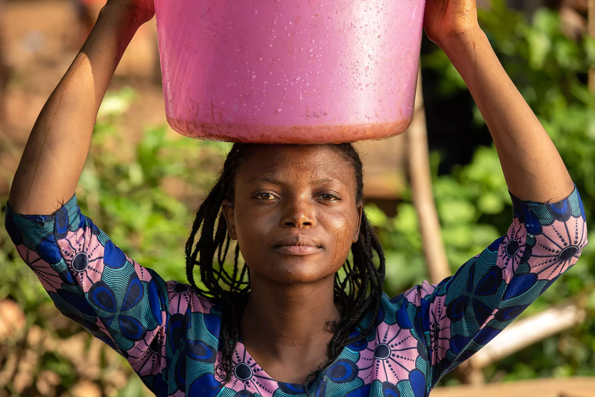 A young woman looks in the camera holding a pink plastic bucket on her head.