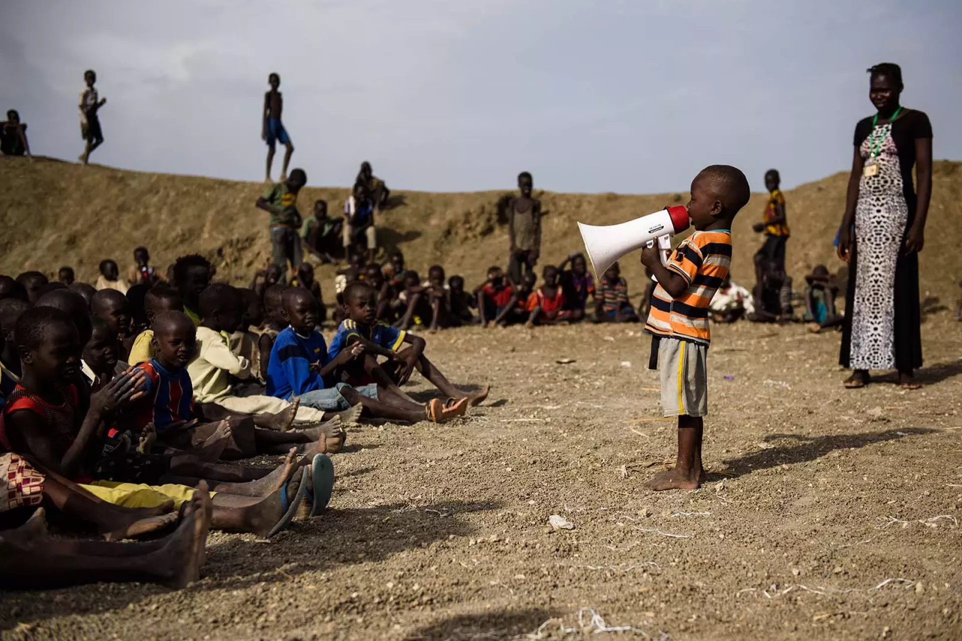 Outdoors, a boy sings into a megaphone for a group of children sitting on the ground and being watched by a woman standing beside them.