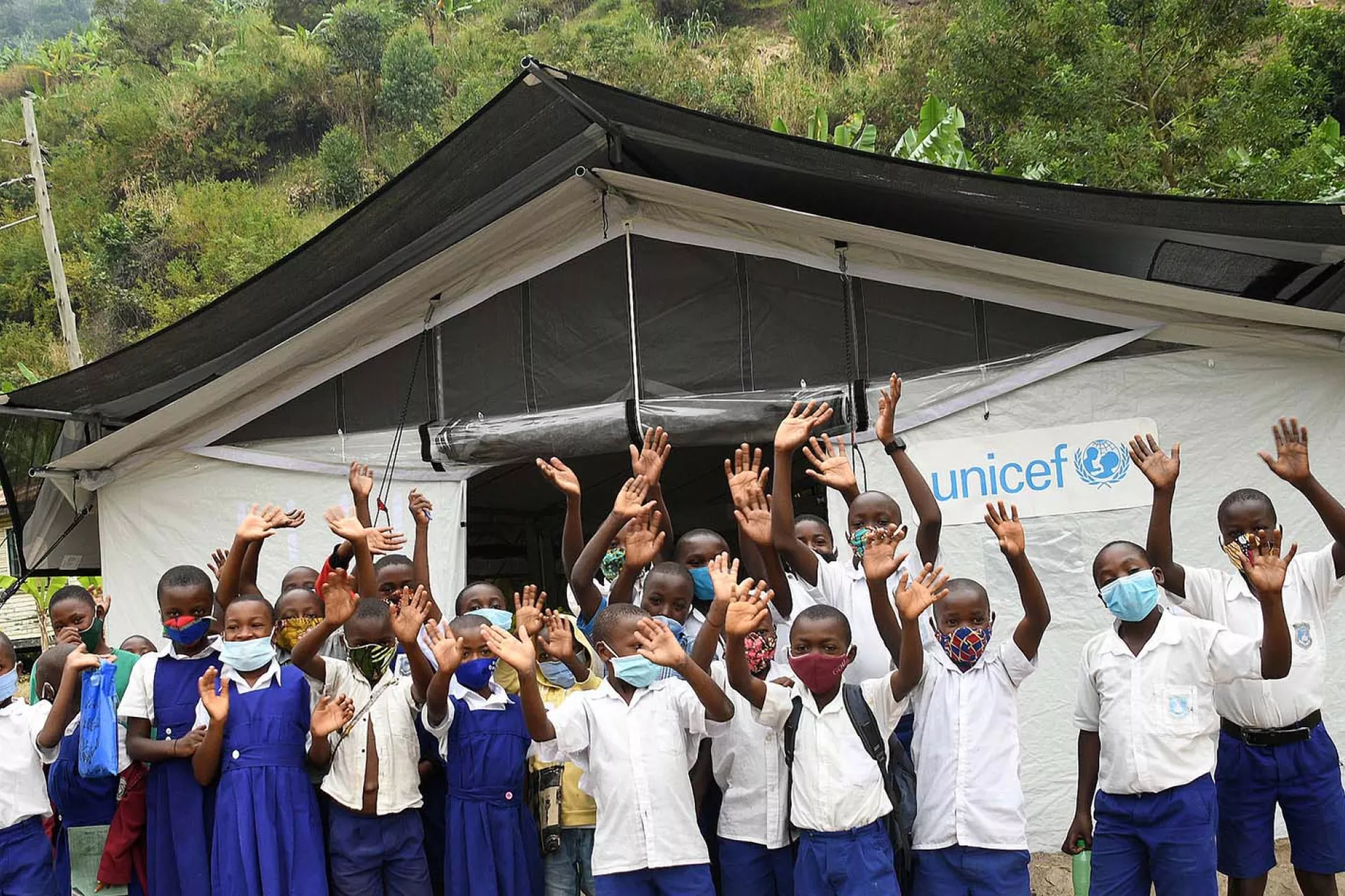 Wearing masks, pupils of a primary school in Kasese District, Uganda, show excitement about the installation of a UNICEF High Performance Tent, which is in the background.