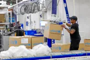 Surrounded by UNICEF-labeled boxes and plastic packaging materials, a worker operates a mechanical arm to lift a cardboard box.