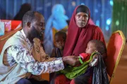Using a MUAC tape, a man screens the arm of a baby held by a woman who looks into the camera while a young child is beside her.