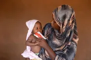 A woman in patterned headscarf feeding an infant with a food sache.