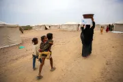 A woman follwed by three children walks in a sandy displacement camp while carrying a large cardbox and a plastic bowl over her head.
