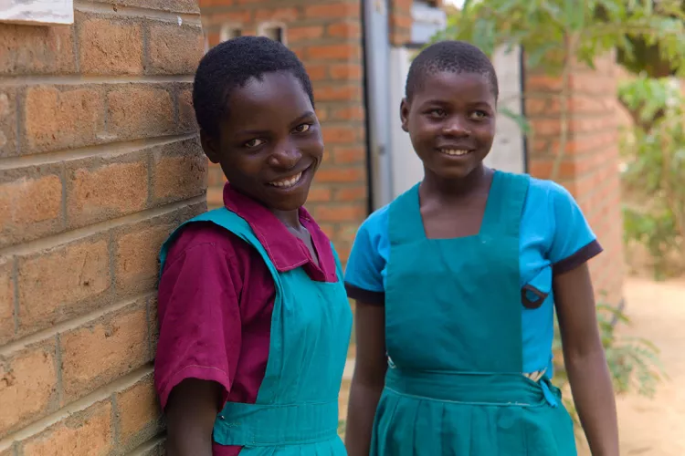 Two girls are smiling in the camera outside of their school. 