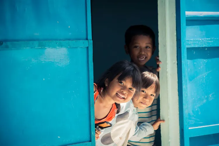 Three children peeking out from behind a partially open bright blue door, with sunlight illuminating their faces.