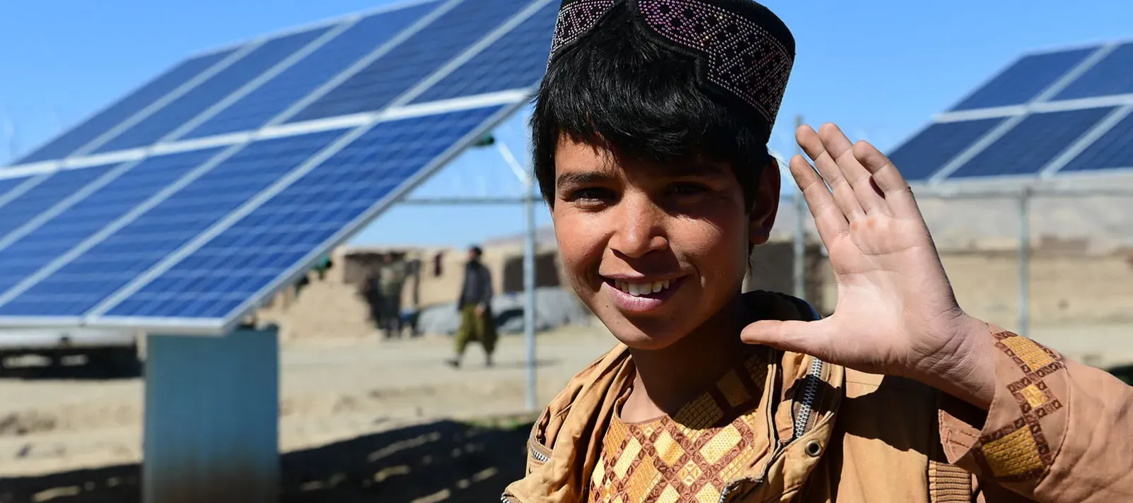A boy waving his left hand with solar panels in the background.