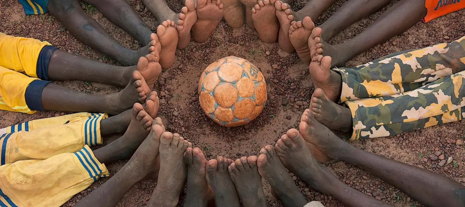 Children&rsquo;s bare feet form a circle surrounding a football on the ground.