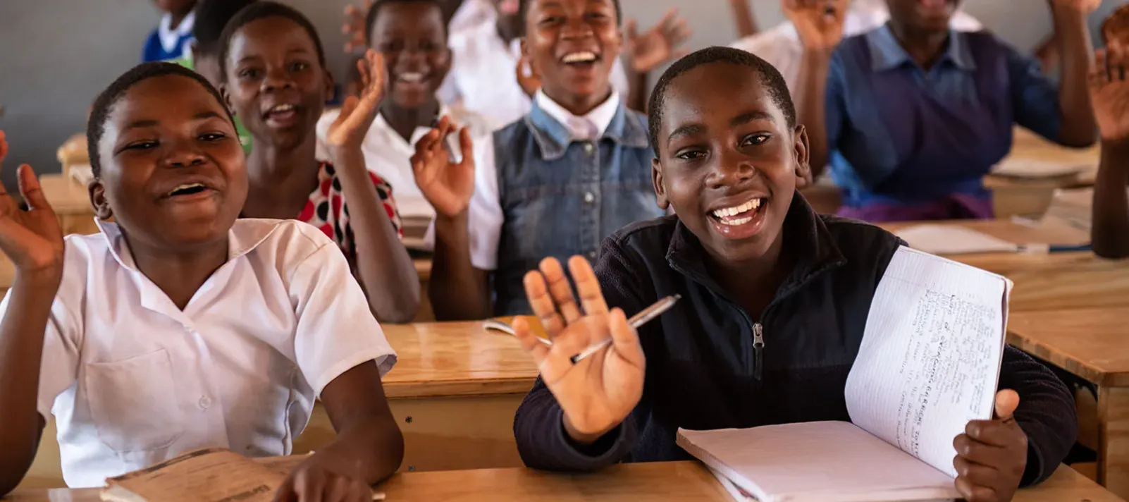 Teenage schoolgirls seated at wooden desks in a classroom, raising their hands and holding notebooks and pens.
