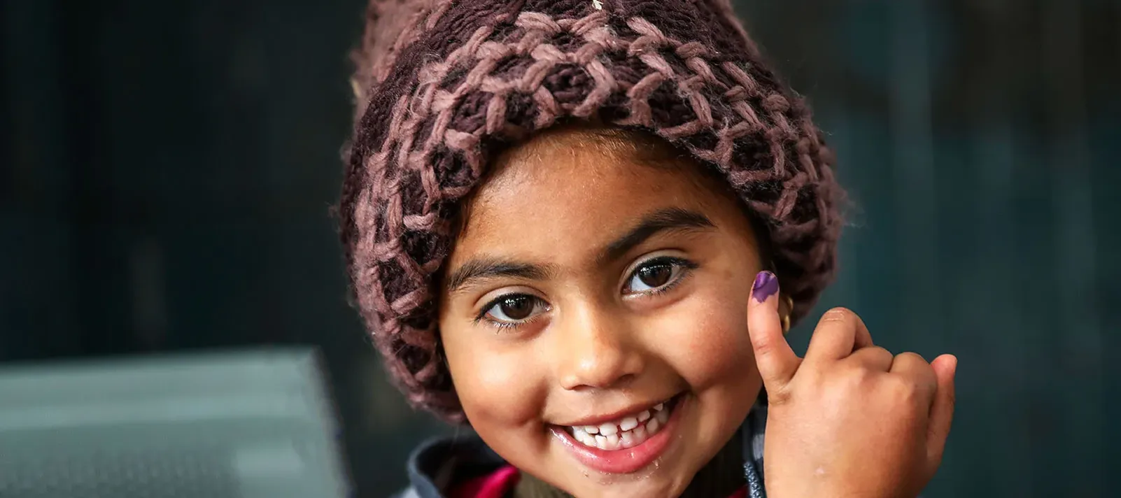 A child-girl smiles while showing her little finger marked with purple ink, which signals that she has just been vaccinated. 