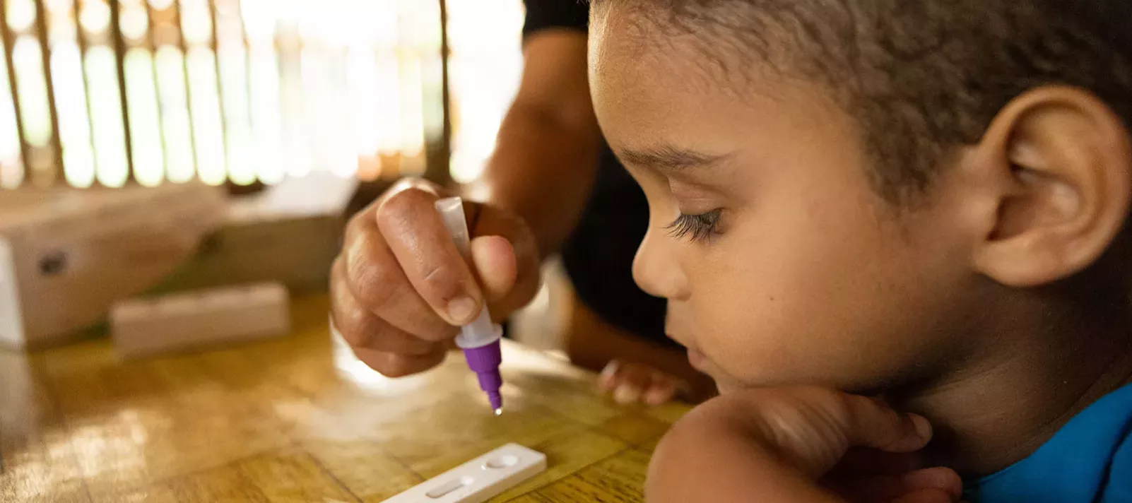 A boy looks at a rapid a diagnostic test while the hand of an adult can be seen manipulating the test. 