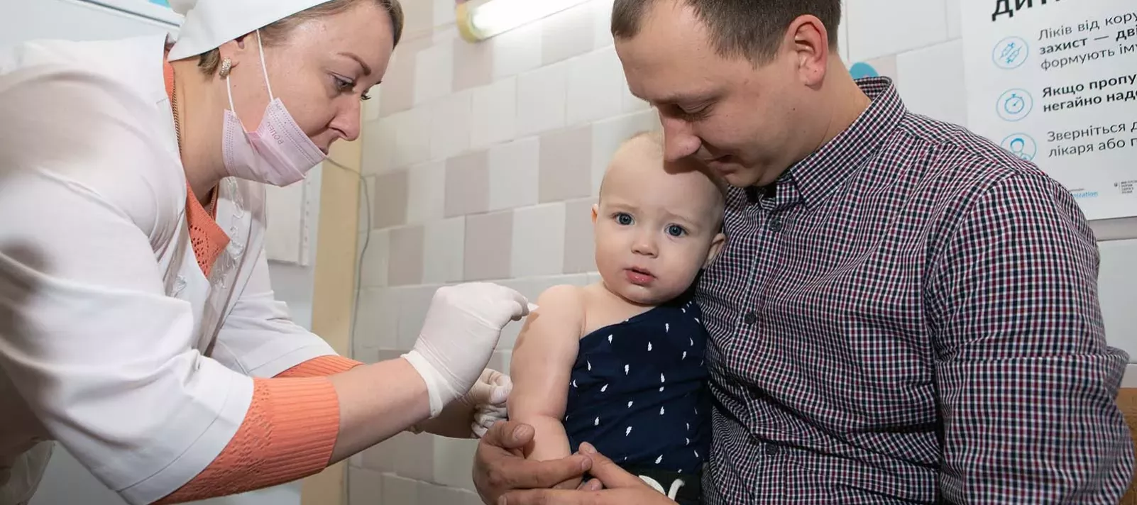 A baby is in the arms of his father is vaccinated by a health care worker. 