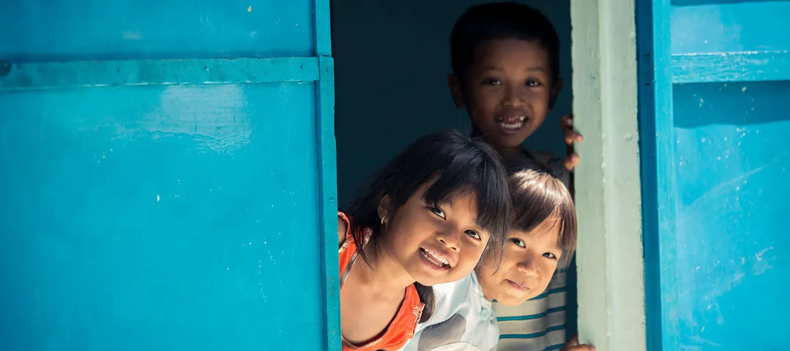 Three children peeking out from behind a partially open bright blue door, with sunlight illuminating their faces.