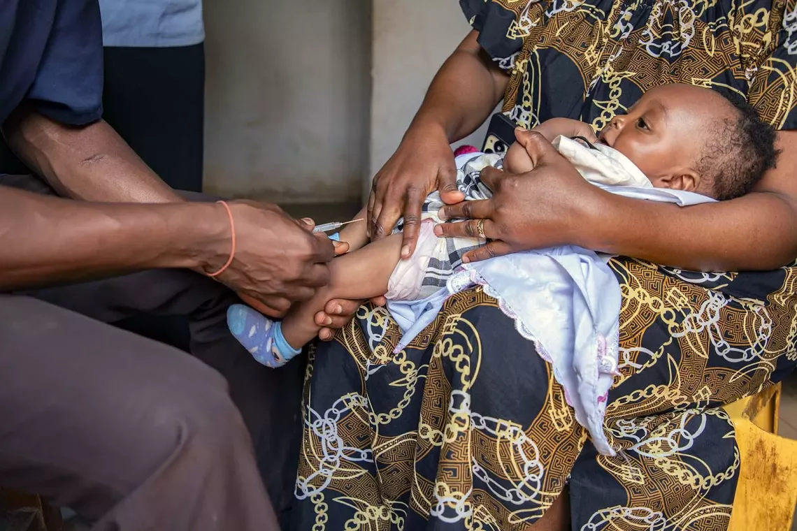 A baby being held by a person is about to receive a shot in her/his left leg. 