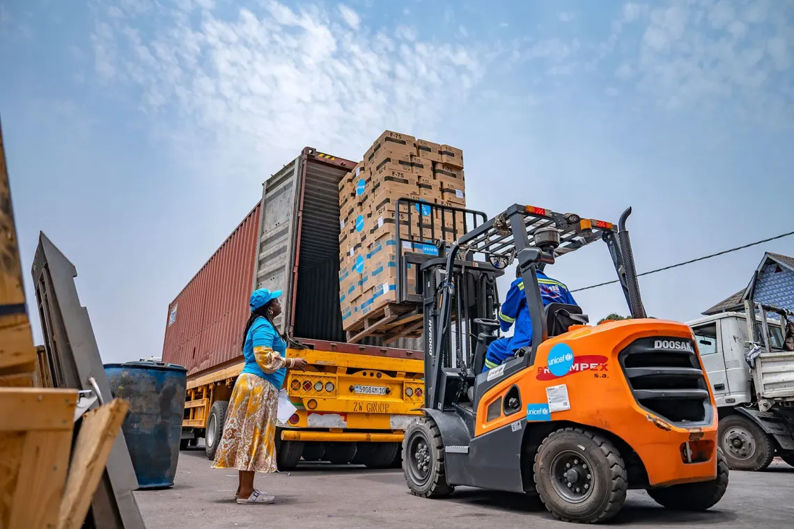 A woman wearing a blue cap stands by a forklift operator unloading stacked boxes from a shipping container onto a truck.