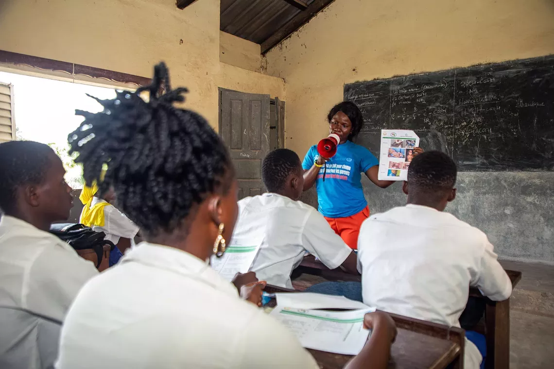 A woman holding a paper sheet talks to a classroom of young students. 