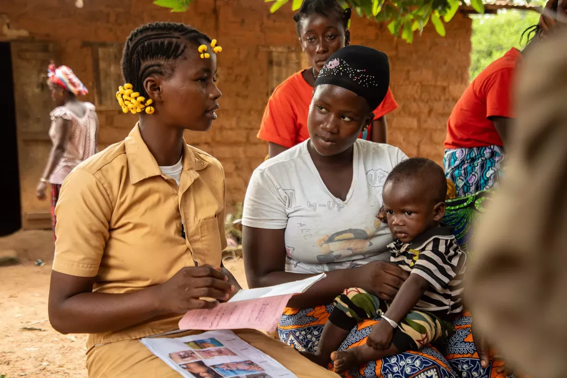 Two young women, one of them with a child in her lap, are sitting and talking in an open air area. 
