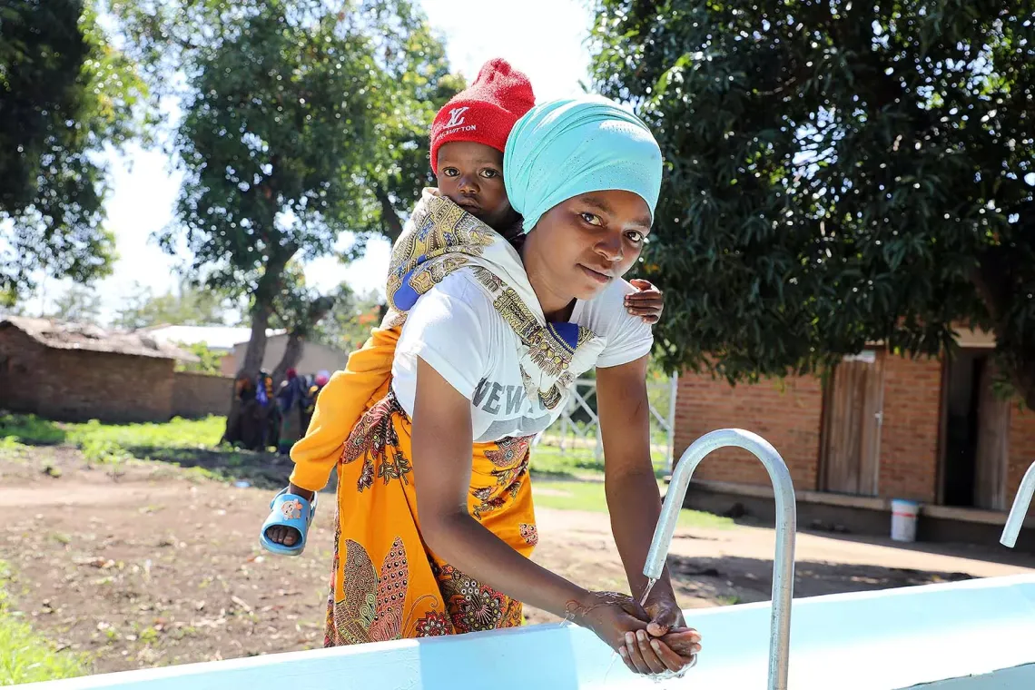 A woman washes her hands at an outdoor sink while carrying a baby on her back. 
