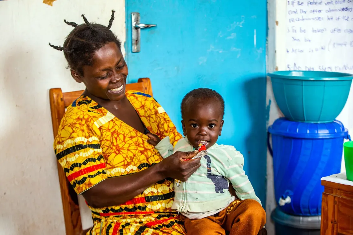 A woman and a child sitting indoors, with the child is eating ready-to-use-therapeutic food (RUTF) from a sachet. 