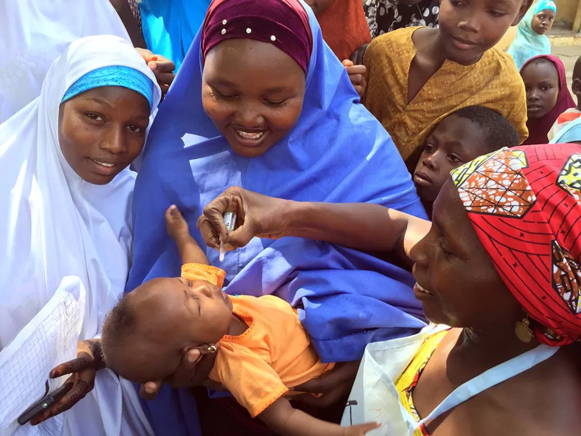 Surrounded by other women and children, a woman holds a baby who is being vaccinated by a health care worker.