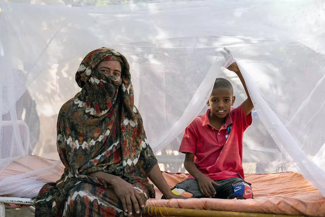 A woman sitting besides a boy who is partially under a mosquito net. 