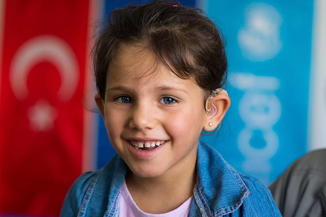 The little girl Sehad, aged 4 years, is wearing a hearing aid and smiling to the camera. 