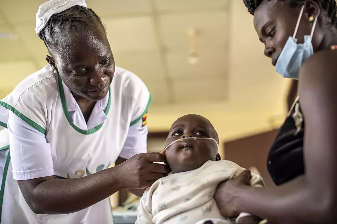 A nurse adjusts a breathing tube in the nose of a baby who sits in the lap of a woman. 