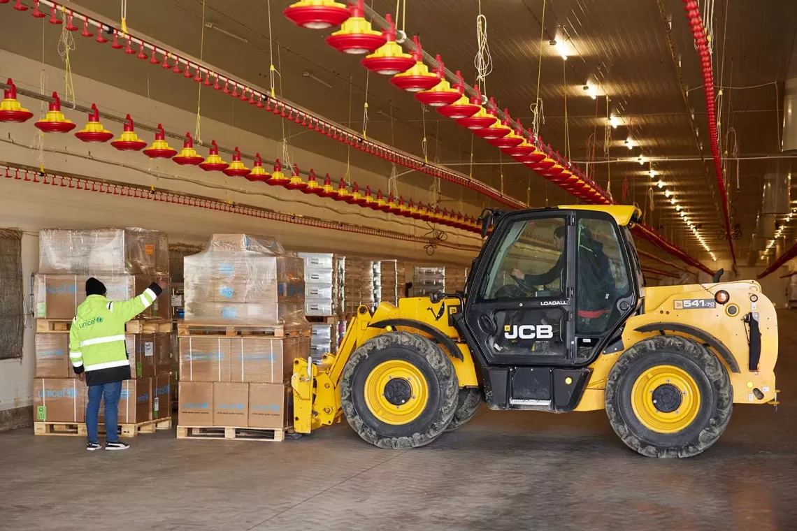 In a warehouse, a UNICEF staff leads another person driving a forklift carrying palettes with cardboard boxes.