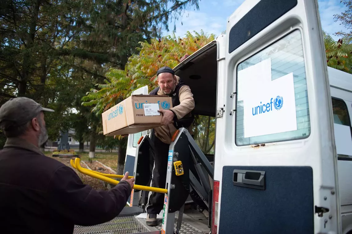 A man on the door of a van hands a cardboard box to another man on the ground. 