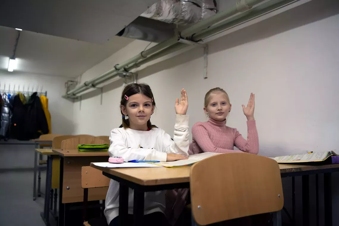 Two sitting at desks in a classroom raise their hands. 