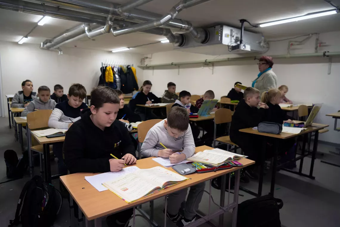  Children sitting at desks and a woman standing among them in a classroom.