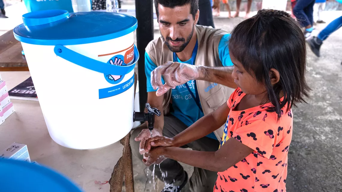 A UNICEF staff shows a girl how to wash hands using water from a white plastic container. 
