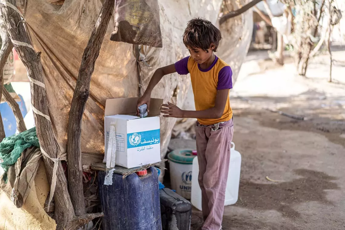 By a makeshift tent, a boy opens a cardboard box displaying the UNICEF logo containing hygiene products.