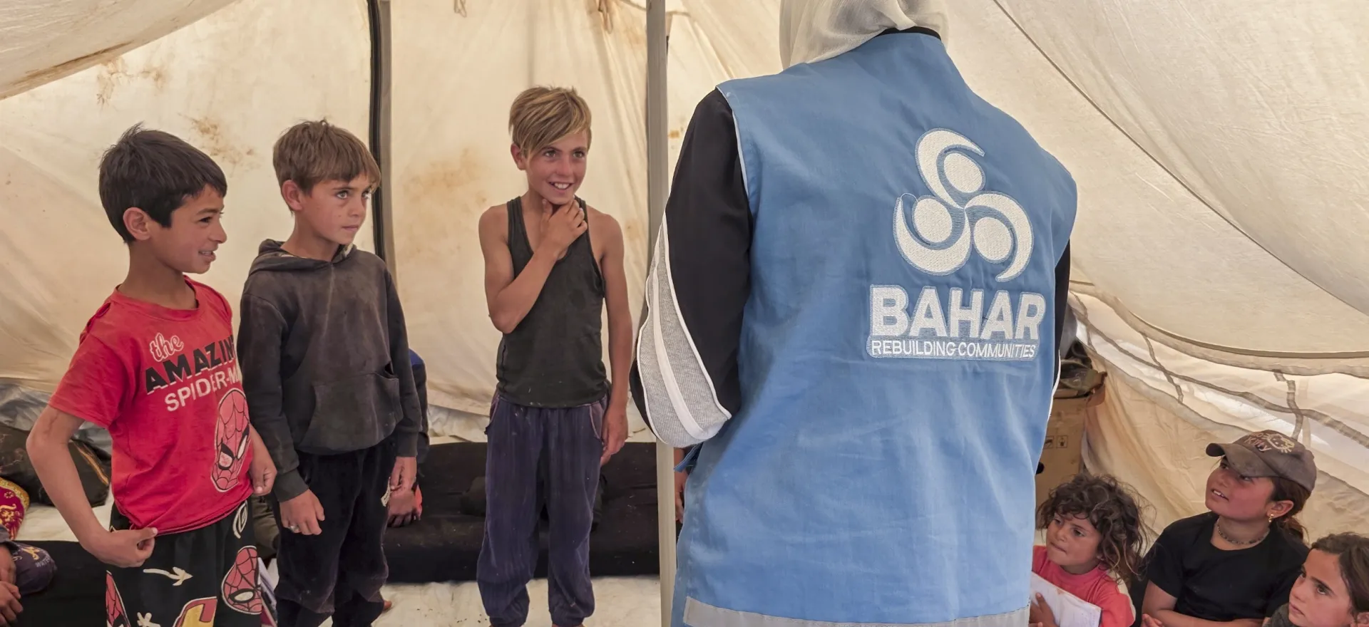 The view from behind a woman wearing a blue jacket and white headscarf at 3 young boys standing next to each other facing her, inside at tent.